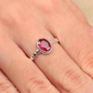 Może przedstawiać: Srebrny pierścionek z dużym owalnym, czerwonym kamieniem szlachetnym. Pierścionek ma wzór z koralików na obrączce w pobliżu kamienia szlachetnego. Pierścionek jest na palcu osoby.