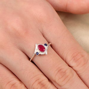 May include: A silver ring featuring a central oval-cut red gemstone, flanked by two smaller blue gemstones. The ring has a decorative, openwork design around the central stone. The ring is displayed on a hand.