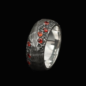 Può includere: Un anello d'argento con una fascia strutturata grigio scuro. L'anello è adornato da diverse piccole gemme rotonde di colore rosso brillante. L'interno dell'anello è in argento lucido. L'anello è su sfondo nero.