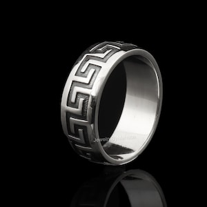 May include: A silver ring with a Greek key pattern around the band. The ring is made of a shiny metal and has a polished finish. The design is a repeating geometric pattern. The ring is displayed against a black background.