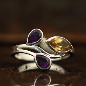 Könnte beinhalten: Ein silberner Ring mit drei Edelsteinen. Zwei tropfenförmige, violette Amethyste flankieren einen blattförmigen, gelben Citrin. Das Design des Rings ist organisch und fließend, mit polierter Oberfläche.