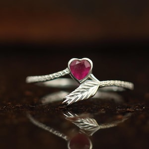 May include: A silver ring featuring a heart-shaped, deep red gemstone. The ring has a leaf design and a textured band. The ring is displayed on a reflective surface, showcasing its intricate details and design.