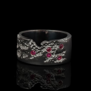 Puede incluir: Un anillo ancho de color gris oscuro con una superficie texturizada y varias piedras preciosas rojas pequeñas y redondas. El anillo tiene un borde orgánico e irregular, y las piedras preciosas están agrupadas en la parte superior.