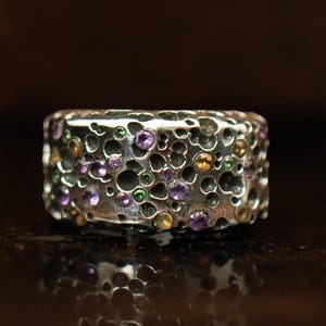 May include: A silver ring with a textured, porous design. The ring is studded with small, colorful gemstones in shades of purple, green, and yellow. The ring's surface reflects the light, creating a shimmering effect.