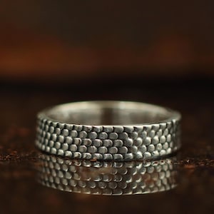 Peut inclure: Une bague en argent avec une surface texturée. La bande de la bague est recouverte d'un motif de petites formes arrondies, lui donnant un aspect tactile unique. La bague est présentée sur une surface réfléchissante.