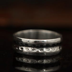Puede incluir: Un anillo de plata con textura martillada. El anillo tiene una banda ancha con una superficie texturizada y una banda más pequeña con un patrón punteado. El anillo se muestra sobre una superficie oscura y reflectante.