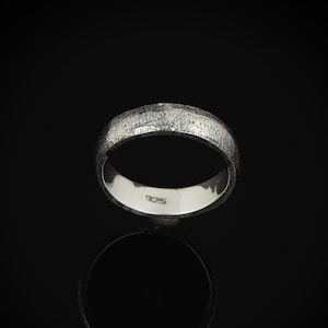 May include: A textured silver ring with a band design. The ring is set against a black background, with a reflection visible below. The inside of the ring is smooth and stamped with "925".