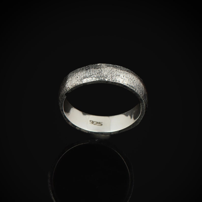 May include: A textured silver ring with a band design. The ring is set against a black background, with a reflection visible below. The inside of the ring is smooth and stamped with "925".