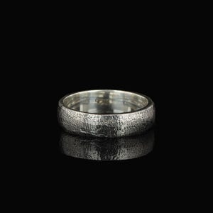 May include: A silver ring with a textured surface. The ring is round and has a band with a rough, hammered appearance. The ring is set against a black background, with its reflection visible below.