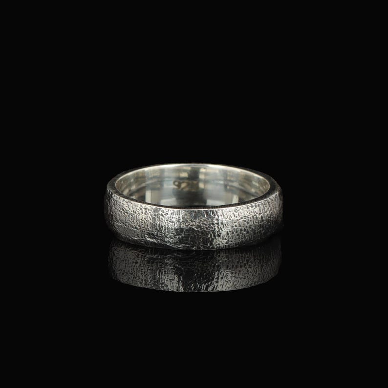 May include: A silver ring with a textured surface. The ring is round and has a band with a rough, hammered appearance. The ring is set against a black background, with its reflection visible below.