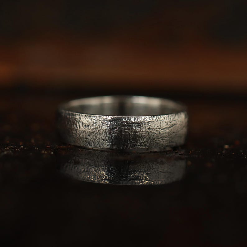 May include: A silver ring with a textured surface. The ring has a band width of approximately 6mm. The ring is displayed on a dark, reflective surface, creating a mirrored effect. The ring's design has a rustic, organic feel.