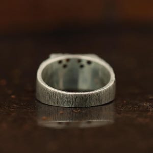 May include: Close-up of a silver ring with a textured surface. The ring has a rectangular top with small, dark red stones. The band is thick and has a brushed metal appearance. The ring is displayed against a blurred, dark background.