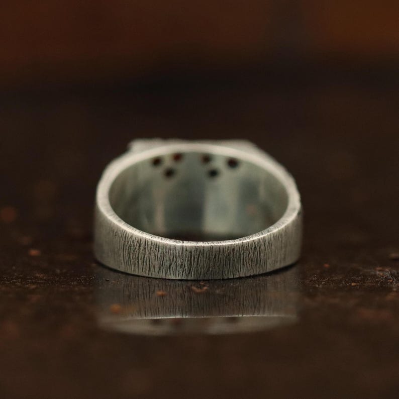 May include: Close-up of a silver ring with a textured surface. The ring has a rectangular top with small, dark red stones. The band is thick and has a brushed metal appearance. The ring is displayed against a blurred, dark background.