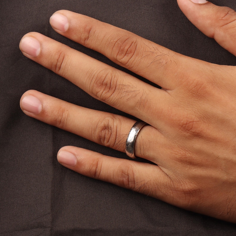 May include: A silver ring with a textured surface is worn on a hand. The ring has a band with a hammered or distressed appearance. The background is a dark, solid color.