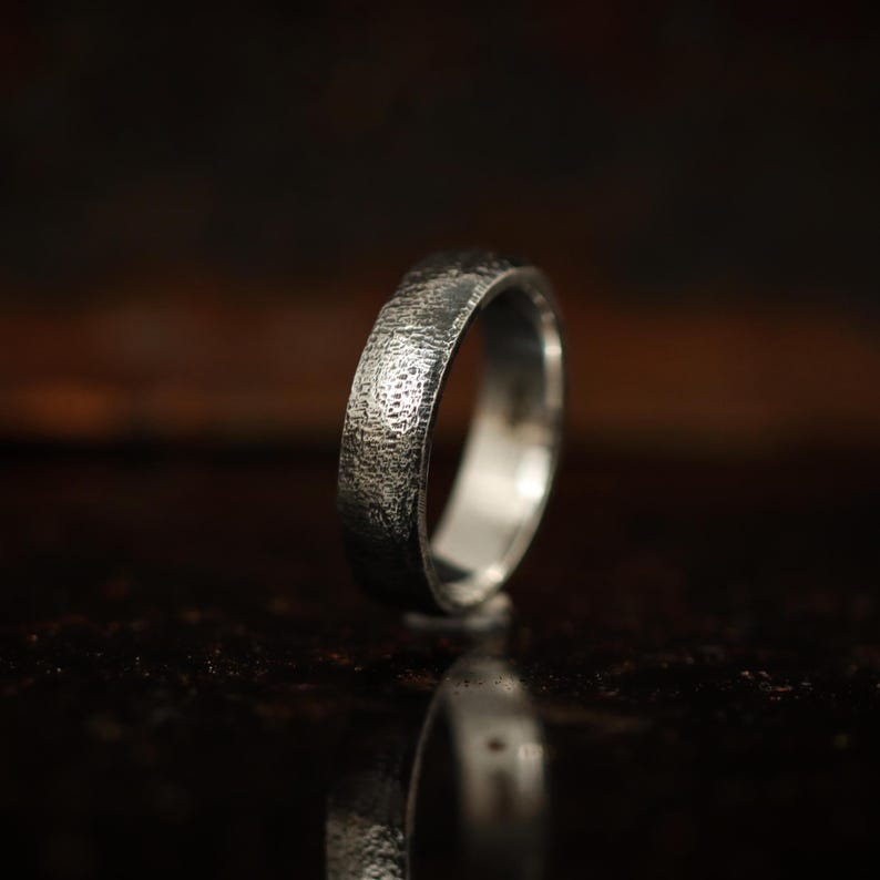 May include: A silver ring with a textured surface. The ring has a hammered or forged appearance, with a band that is approximately 0.2 inches wide. The ring is displayed on a reflective surface, showcasing its polished interior.