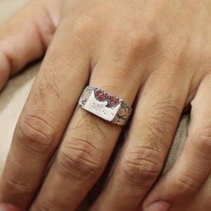 May include: A silver-toned ring with a rectangular face and a textured surface. The ring is adorned with small, round, red gemstones. The design has a rustic, handcrafted appearance, suitable for everyday wear.