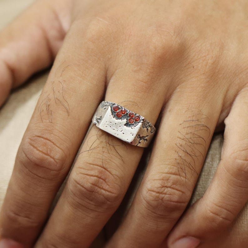 May include: A silver-toned ring with a rectangular face and a textured surface. The ring is adorned with small, round, red gemstones. The design has a rustic, handcrafted appearance, suitable for everyday wear.
