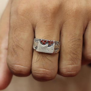 May include: A silver ring with a rectangular face and a textured band. The face of the ring features a cluster of small, round, red gemstones. The ring is worn on a finger, showcasing its design and craftsmanship.