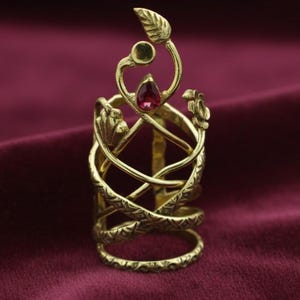 Può includere: Un anello color oro con un design intricato con foglie, un fiore e una gemma rossa a forma di goccia. L'anello ha una struttura unica e stratificata con fasce intrecciate.