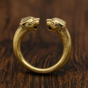 Könnte beinhalten: Ein goldfarbener Ring mit zwei stilisierten Löwenköpfen, die sich gegenüberstehen. Der Ring hat ein kreisförmiges Band und besteht aus einem glänzenden, metallischen Material. Die Löwenköpfe weisen detaillierte Merkmale auf, darunter offene Münder und sichtbare Augen.
