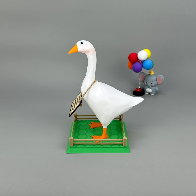 May include: A white goose ornament with an orange beak and feet stands on a green base with a small fence. A small elephant ornament with balloons is in the background. The goose has a tag around its neck.