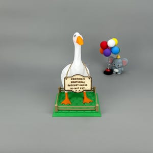 May include: A novelty ornament featuring a white goose with orange feet and beak, holding a sign that reads "CRISTINA'S EMOTIONAL SUPPORT GOOSE. DO NOT PET." An elephant figurine with balloons is also visible.