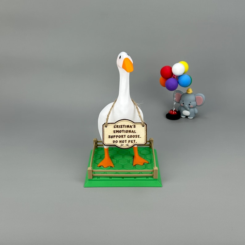 May include: A novelty ornament featuring a white goose with orange feet and beak, holding a sign that reads "CRISTINA'S EMOTIONAL SUPPORT GOOSE. DO NOT PET." An elephant figurine with balloons is also visible.