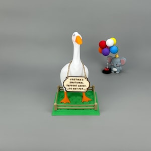 May include: A white goose ornament with orange feet and beak, holding a wooden sign that reads "Cristina's Emotional Support Goose. DO NOT PET." A small elephant figurine with balloons is to the right.
