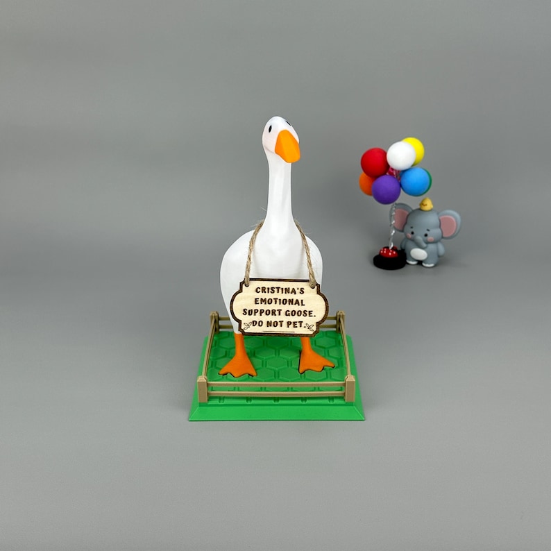 May include: A white goose ornament with orange feet and beak, holding a wooden sign that reads "Cristina's Emotional Support Goose. DO NOT PET." A small elephant figurine with balloons is to the right.