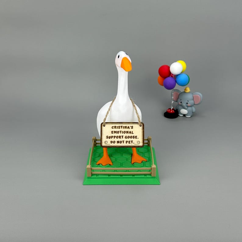May include: A white goose ornament with an orange beak and feet, holding a sign that reads "Cristina's Emotional Support Goose. Do Not Pet." A small elephant ornament with balloons is to the right. The goose stands on a green base.