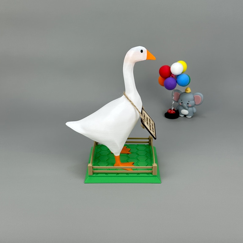 May include: A white goose ornament with orange feet and beak stands on a green base with a small fence. A small grey elephant ornament holds colourful balloons. The goose has a tag hanging from its neck.