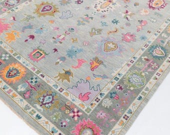 Può includere: Un tappeto rettangolare con sfondo azzurro e un motivo floreale dettagliato. Il tappeto presenta un bordo con disegni floreali e geometrici colorati in rosa, arancione, verde e viola. Il design centrale include varie forme e colori.