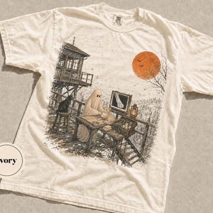 Puede incluir: Una camiseta de manga corta color marfil con un estampado gráfico detallado de un fantasma sentado en un escritorio con un gato, un monitor que muestra una radiografía, una linterna y una torre de vigilancia bajo una gran luna naranja.