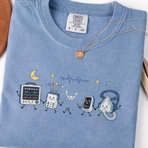 Könnte beinhalten: Ein blaues T-Shirt mit einem Grafikdruck von fünf anthropomorphen medizinischen Geräten, die sich an den Händen halten. Das Design zeigt einen Herzmonitor, ein EKG-Gerät, ein Stethoskop, ein Pulsoximeter und eine CPAP-Maske mit einem Halbmond.