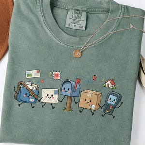 Op de afbeelding: Een mosgroen t-shirt met ronde hals en een grafische print van een lachende posttas, envelop, brievenbus, kartonnen doos en handscanner die elkaars hand vasthouden. Het shirt heeft een Comfort Colors label in de kraag.