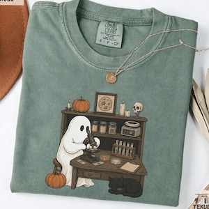 Könnte beinhalten: Ein moosgrünes Kurzarm-T-Shirt mit einer Grafik eines weißen Geistes, der an einem Holztisch sitzt und ein Mikroskop benutzt. Auf dem Tisch stehen Reagenzgläser, Gläser und ein Kürbis, während eine schwarze Katze darunter schläft.