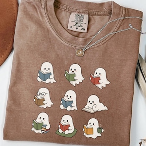 Könnte beinhalten: Ein espressofarbenes T-Shirt mit einem Grafikdruck von neun kleinen weißen Geistern, die Bücher lesen. Das T-Shirt hat einen Rundhalsausschnitt und ein Comfort Colors Markenetikett im Kragen. Eine silberne Halskette liegt auf dem Stoff.