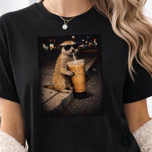 May include: Black t-shirt with a graphic of a meerkat wearing sunglasses and drinking an iced coffee. The meerkat is on a curb at night. The word "Black" is in the upper left corner.