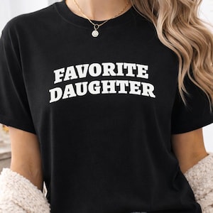 以下が含まれることがあります： 「FAVORITE DAUGHTER」の白く太字の文字がプリントされた黒いTシャツ。半袖のシャツは柔らかい素材で作られているようです。モデルは金のネックレスとイヤリングを着用しています。