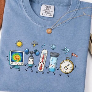 Könnte beinhalten: Ein blaues T-Shirt mit einer Grafik von fünf anthropomorphen Wetterinstrumenten, die sich an den Händen halten, darunter ein Radarschirm, ein Anemometer, ein Thermometer, ein Regenmesser und ein Barometer, sowie Sonnen- und Wolkensymbole.
