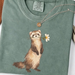Könnte beinhalten: Ein moosgrünes T-Shirt mit Rundhalsausschnitt und einer grafischen Illustration eines stehenden Frettchens, das ein weißes Gänseblümchen im Maul hält. Das Shirt hat ein Comfort Colors Markenetikett am Innenhals mit Größe S-P-CH und Made in Honduras.
