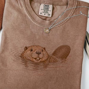 May include: An espresso-colored short-sleeve t-shirt featuring a detailed line-art illustration of a beaver swimming in water. The shirt has a crew neckline and a Comfort Colors brand label on the inside collar. A silver chain necklace rests on top.