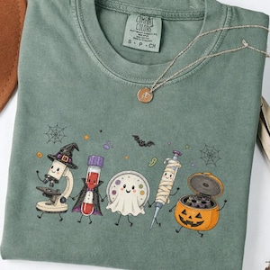 Peut inclure: Un t-shirt à manches courtes vert mousse avec une impression graphique représentant un microscope de dessin animé portant un chapeau de sorcière, un tube à essai vampire, un fantôme, une seringue momie et une citrouille d'Halloween.