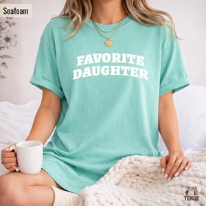 以下が含まれることがあります： 「FAVORITE DAUGHTER」の白文字がプリントされた、シーフォームグリーンのTシャツ。半袖でゆったりとしたフィット感。白いマグカップが左手に持たれています。モデルは金のネックレスを着用しています。