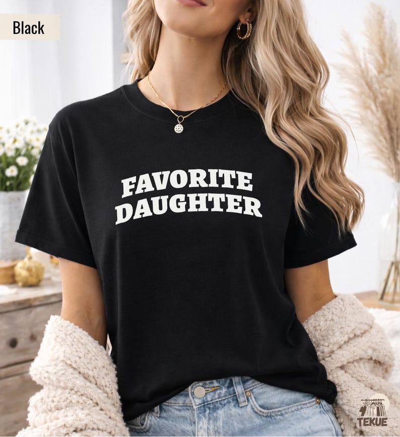 以下が含まれることがあります： 「FAVORITE DAUGHTER」の白く太字の文字がプリントされた黒いTシャツ。半袖のシャツは柔らかい素材で作られているようです。モデルは金のネックレスとイヤリングを着用しています。