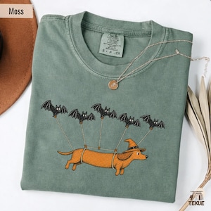Könnte beinhalten: Ein moosgrünes T-Shirt mit Rundhalsausschnitt und einem Aufdruck eines braunen Dackels mit Hexenhut, der von fünf schwarzen Fledermäusen an Seilen in der Luft gehalten wird. Das Etikett am Innenkragen zeigt die Marke Comfort Colors und Größe S P CH.