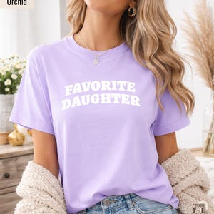 以下が含まれることがあります： 「FAVORITE DAUGHTER」の白文字がプリントされた、薄紫色のTシャツ。クルーネックのシャツは無地です。モデルは青いジーンズとクリーム色のカーディガンを着ています。「Orchid」の文字が左上にあります。