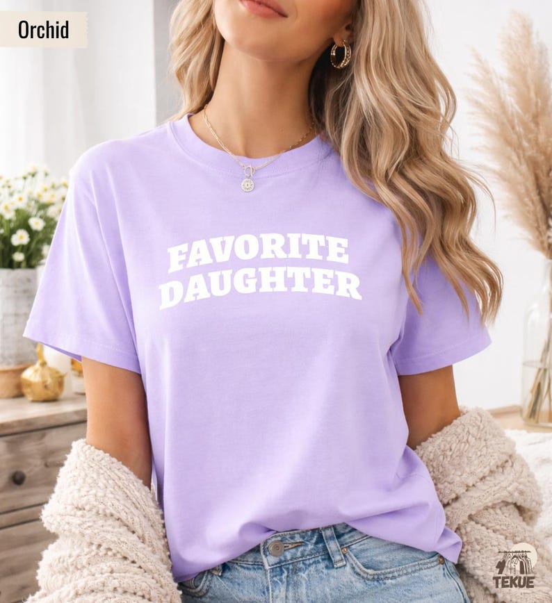 以下が含まれることがあります： 「FAVORITE DAUGHTER」の白文字がプリントされた、薄紫色のTシャツ。クルーネックのシャツは無地です。モデルは青いジーンズとクリーム色のカーディガンを着ています。「Orchid」の文字が左上にあります。
