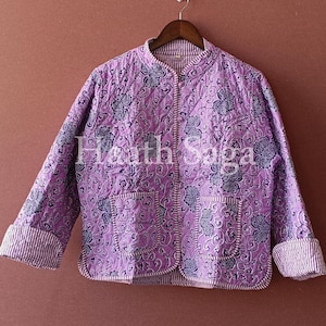 May include: A quilted jacket in shades of lavender and gray, featuring a floral pattern. The jacket has a round collar, two front pockets, and striped cuffs. The text "Hearth Saga" is visible on the jacket.