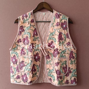 Può includere: Un gilet trapuntato con un motivo floreale nei toni del pesca, viola e verde. Il gilet ha due tasche e una fodera a righe. È appeso a una gruccia di legno su uno sfondo marrone.